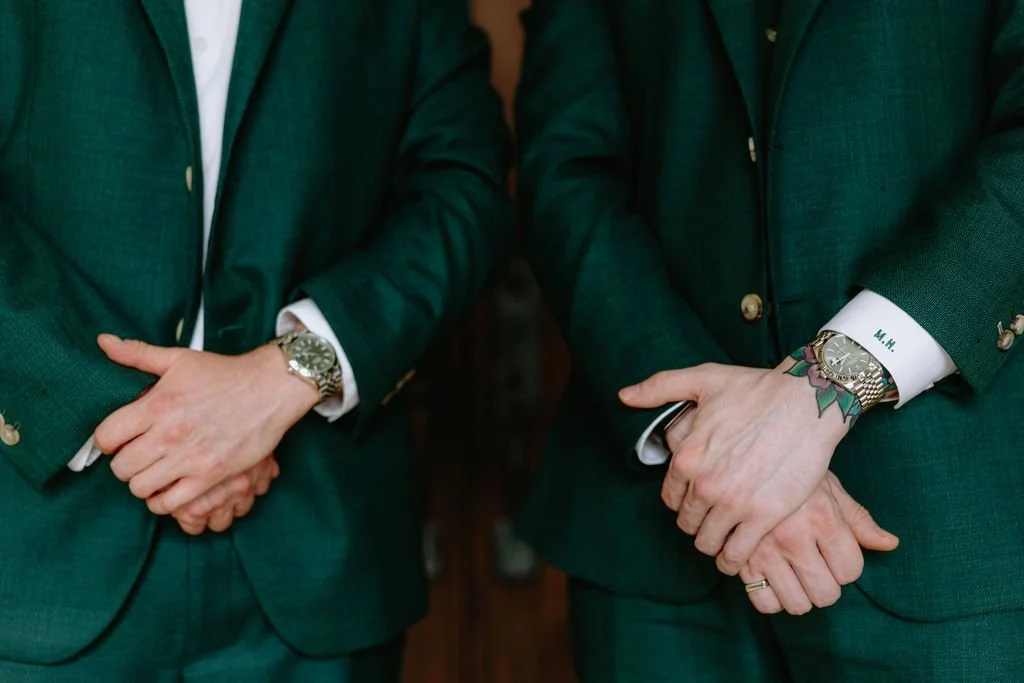 Two people dressed in green suits with white shirts, standing close and holding hands with interlocked fingers, showing watches and a tattoo on one person's wrist.