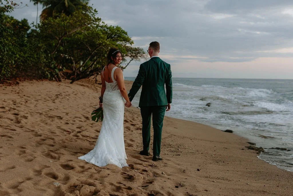 A bride and groom in wedding attire walking on the beach holding hands, with greenery to the left and ocean waves in the background.