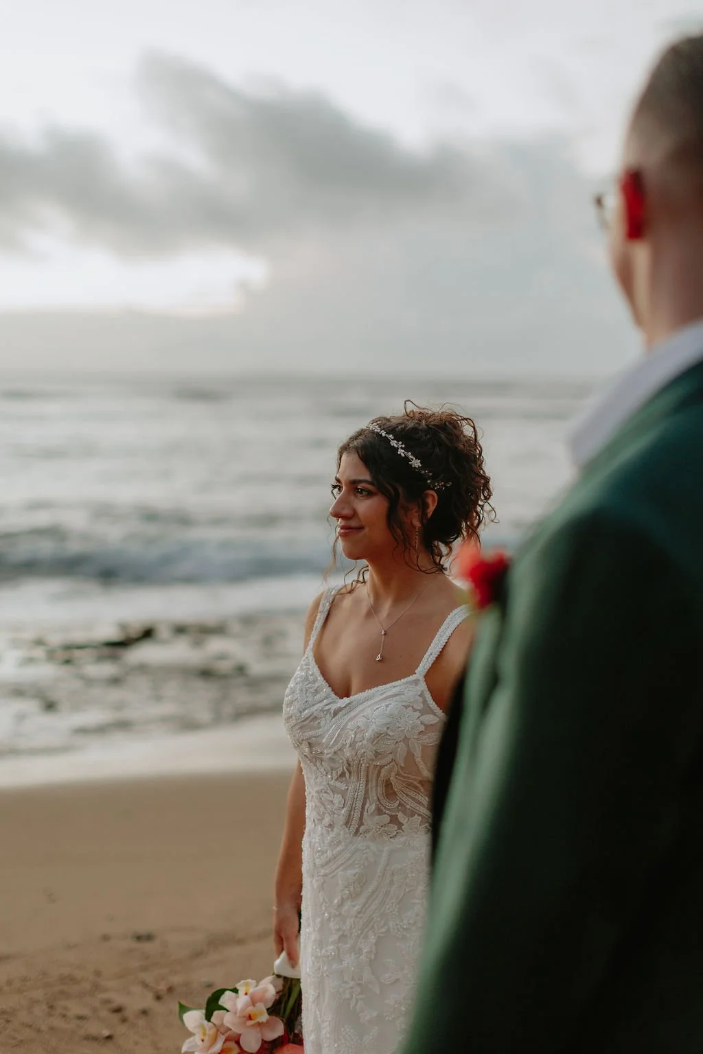 A bride in a white lace wedding dress holding a bouquet of pink and white flowers, standing on a beach with the ocean and cloudy sky in the background, during her wedding ceremony.