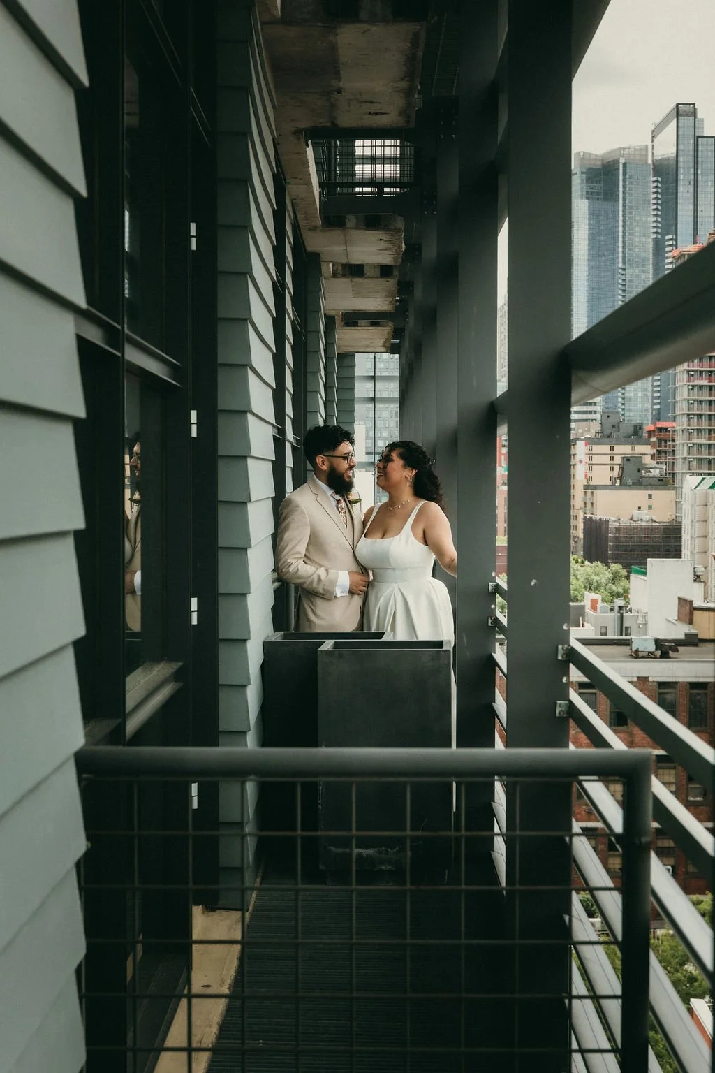 A couple dressed in wedding attire, standing close and smiling at each other on a high-rise balcony in an urban cityscape.