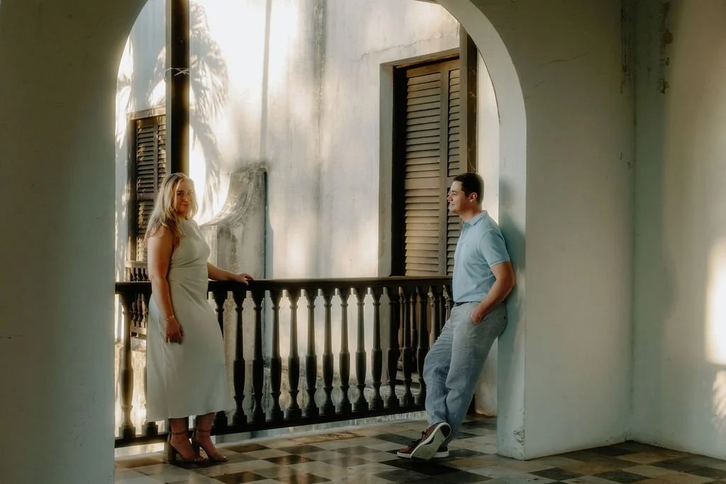 A woman in a white dress and high heels standing on a balcony, facing a man in a light blue shirt and gray pants who is leaning against a wall, in a sunlit room with a checkered floor.