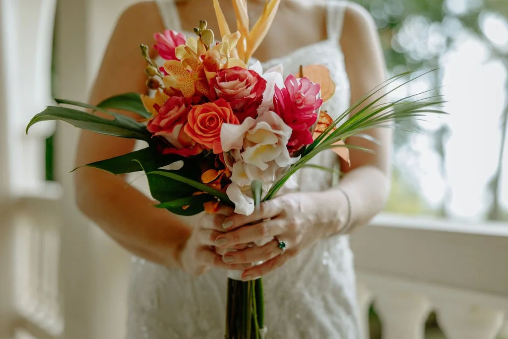 A woman in a white dress holding a colorful bouquet of flowers with pink, orange, yellow, and white blooms, and green leaves, standing indoors with sunlight coming through the windows.