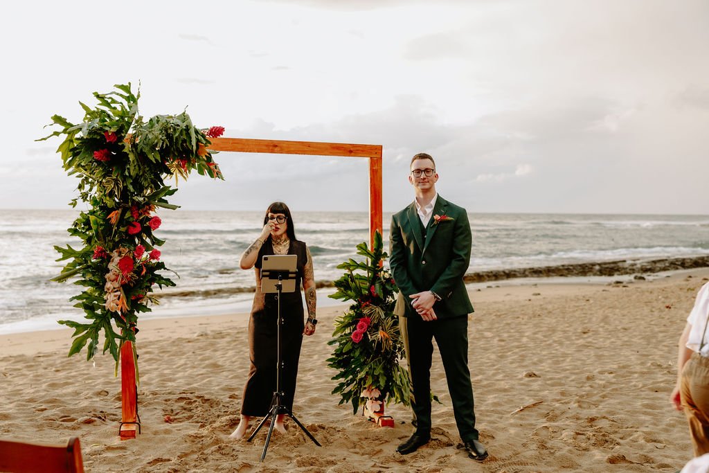 A wedding ceremony taking place on the beach with a man in a green suit standing under a wooden arch decorated with flowers, and a woman with tattoos in a dress reading from a tablet nearby.