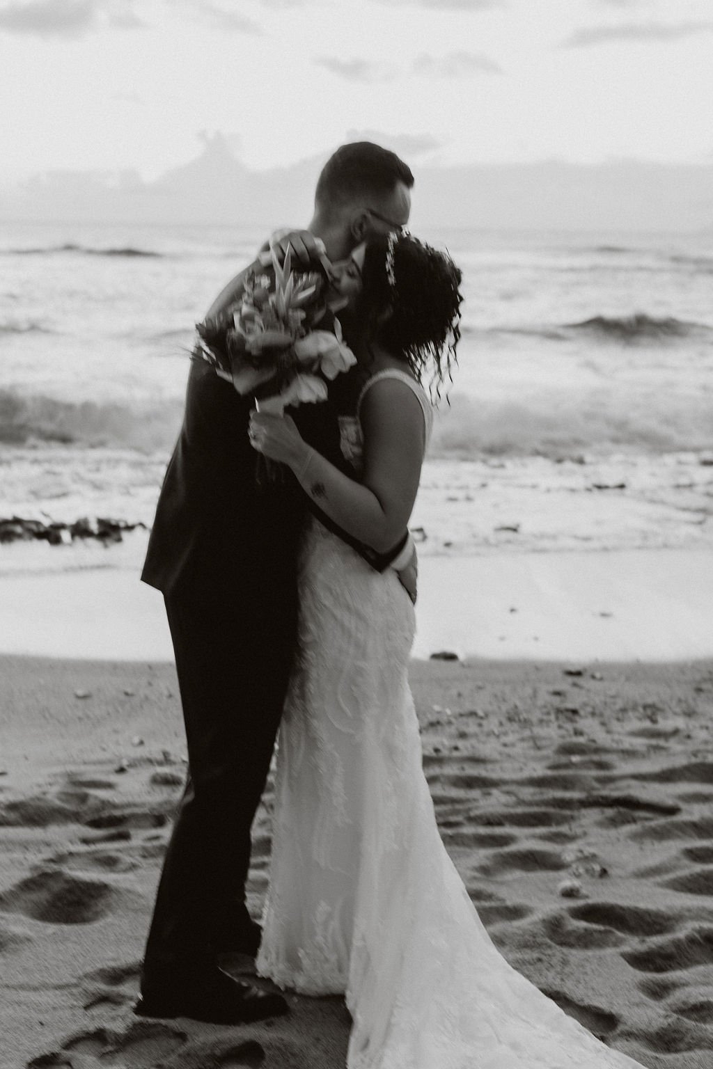 A couple dressed in wedding attire embracing on a beach, the man holding a bouquet of flowers, with ocean waves in the background.