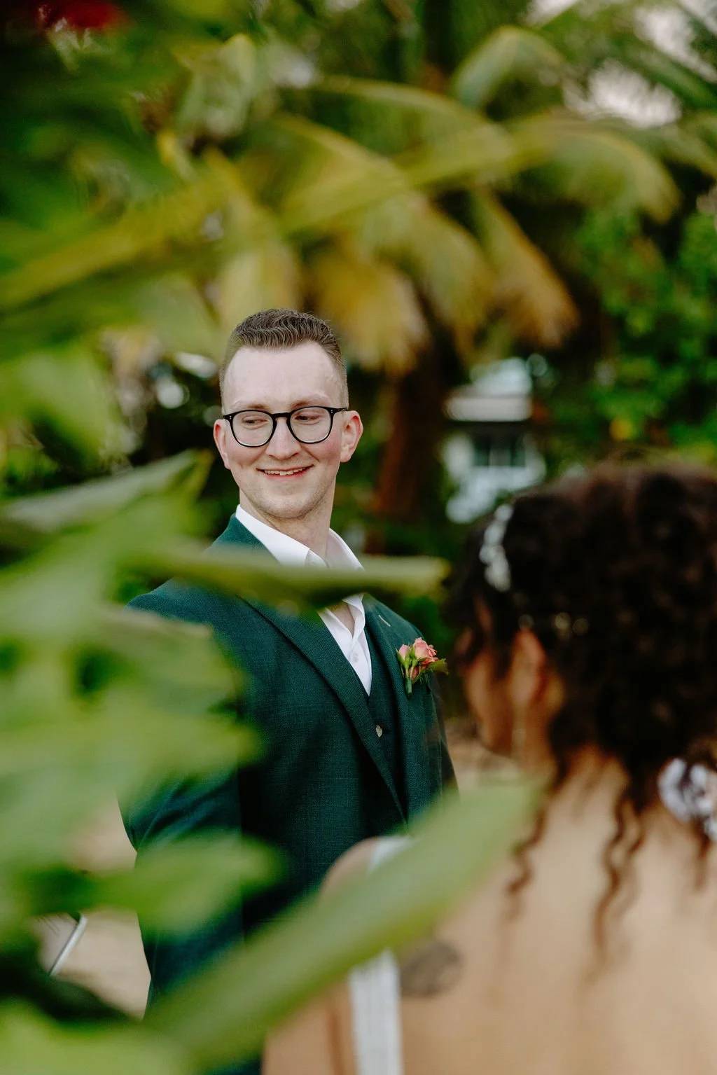 A groom in a suit with glasses and a pink flower boutonniere, smiling at a bride with curly dark hair and a white dress, outdoors with green foliage and trees in the background.