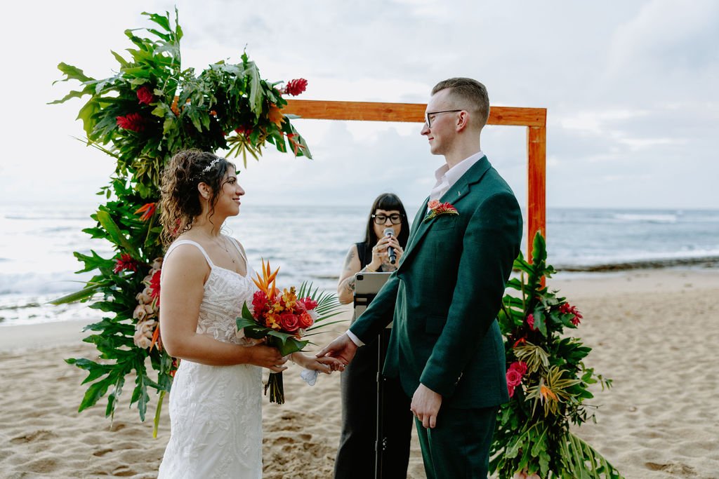 A couple getting married on the beach, holding hands, exchanging rings, with an officiant in the background, and tropical floral arrangements framing their ceremony.
