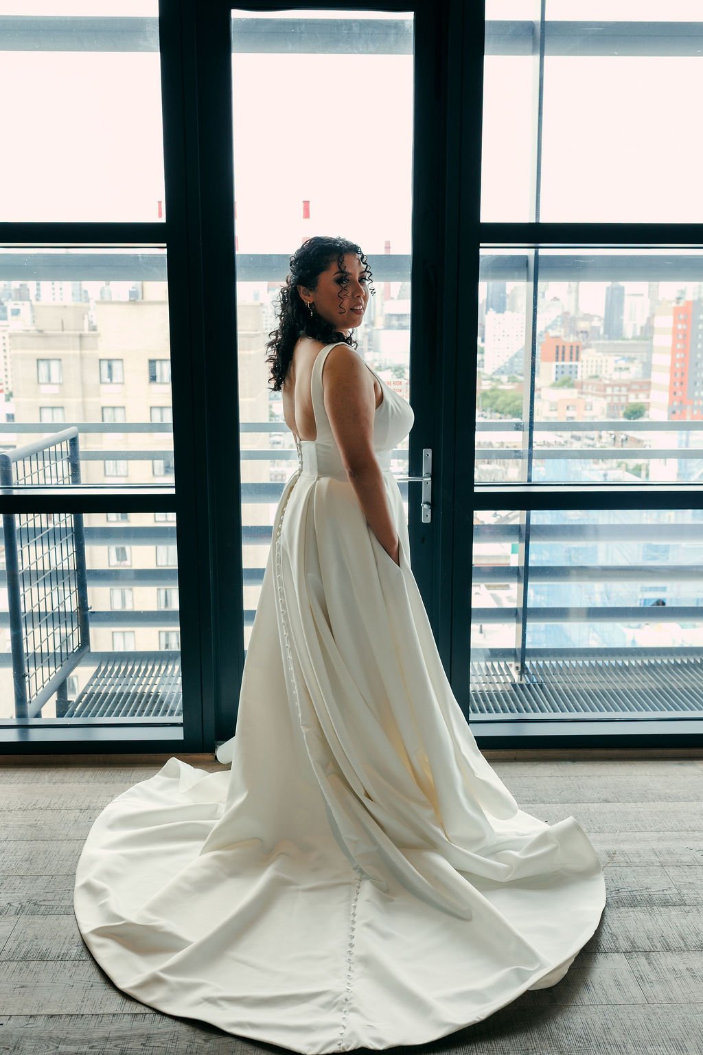 A woman in a white wedding dress standing indoors in front of large glass doors overlooking a city skyline.