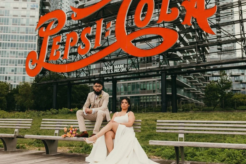 A man in a beige suit and a woman in a white wedding dress sitting on a bench in front of a large Pepsi-Cola sign in an urban park area.
