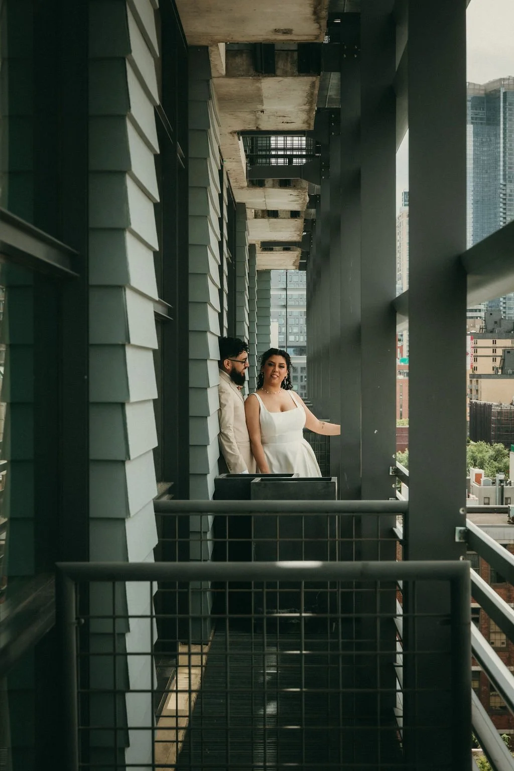 A man and woman standing on a balcony of a high-rise building in an urban area, with city buildings in the background. The woman is wearing a white dress and the man is wearing a light-colored suit.