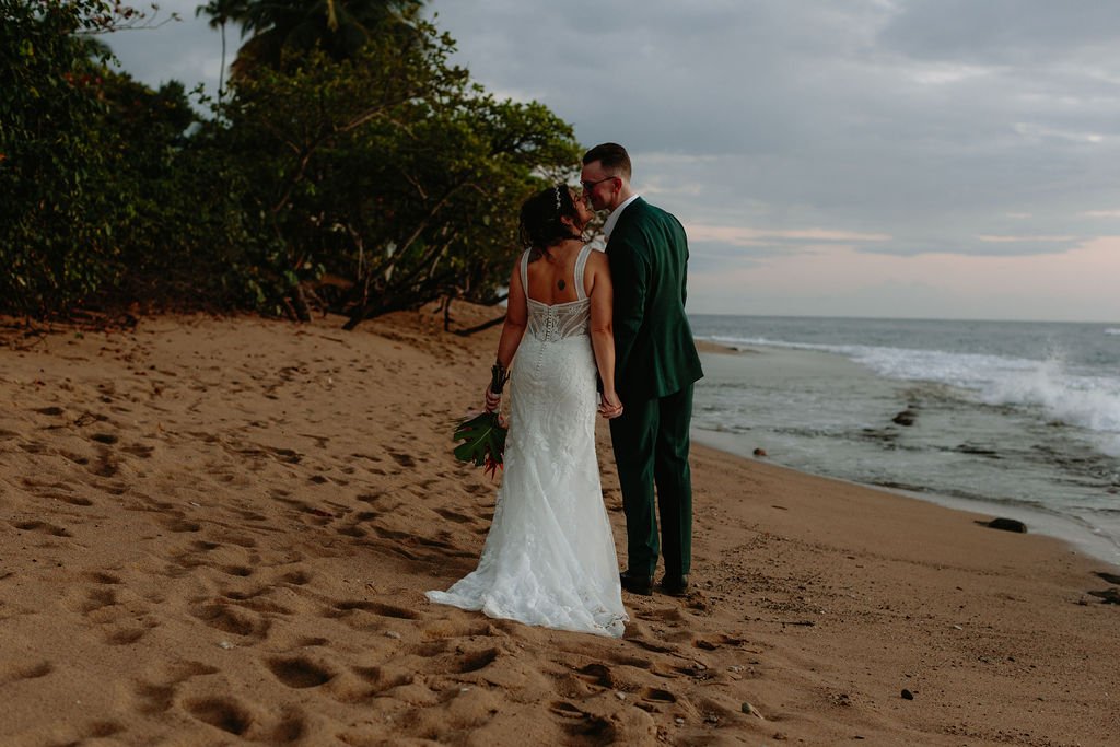 A bride and groom in wedding attire sharing a moment on a sandy beach at sunset with waves in the background.