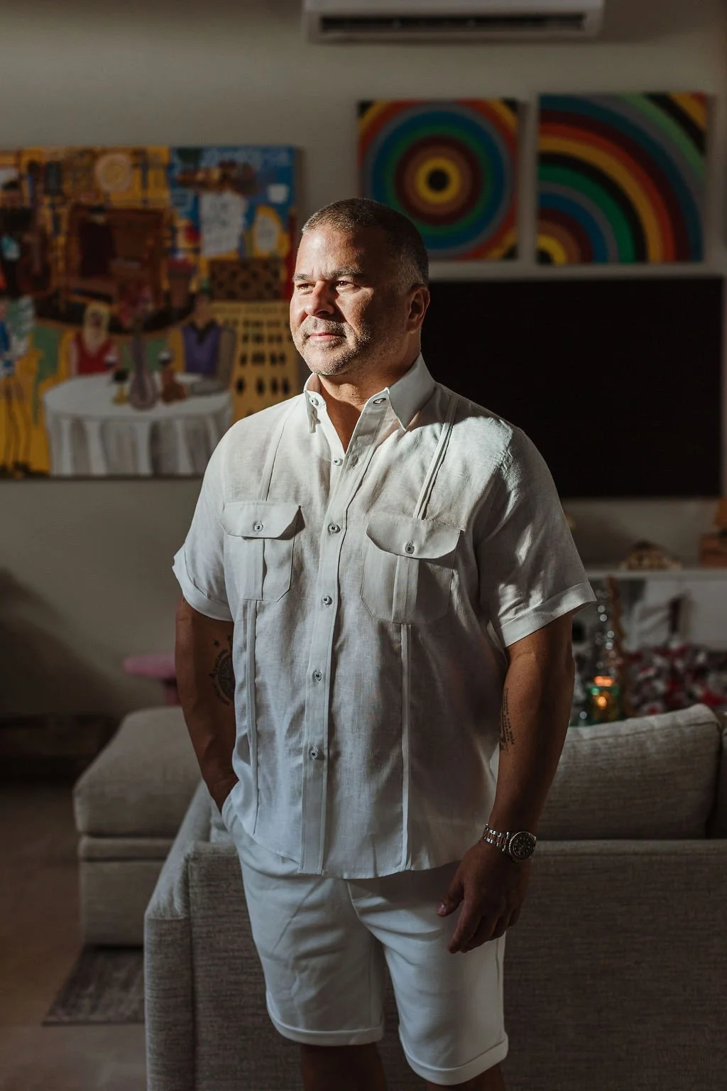 A man in white shirt and shorts standing in a living room, illuminated by sunlight, with colorful abstract paintings on the wall behind him.