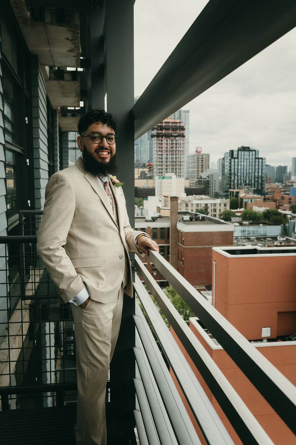 A man in a beige suit, glasses, and a boutonniere standing on a balcony with a cityscape in the background.