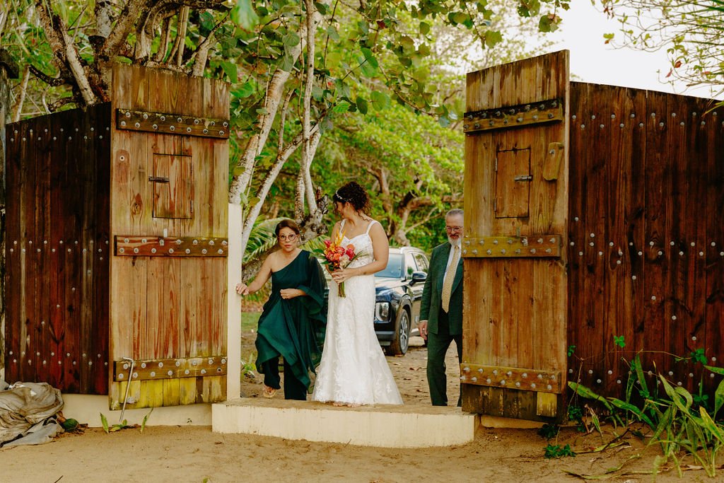 A bride in a white dress holding a bouquet, walking through an open wooden gate with two people on either side, in an outdoor setting with trees and parked cars in the background.