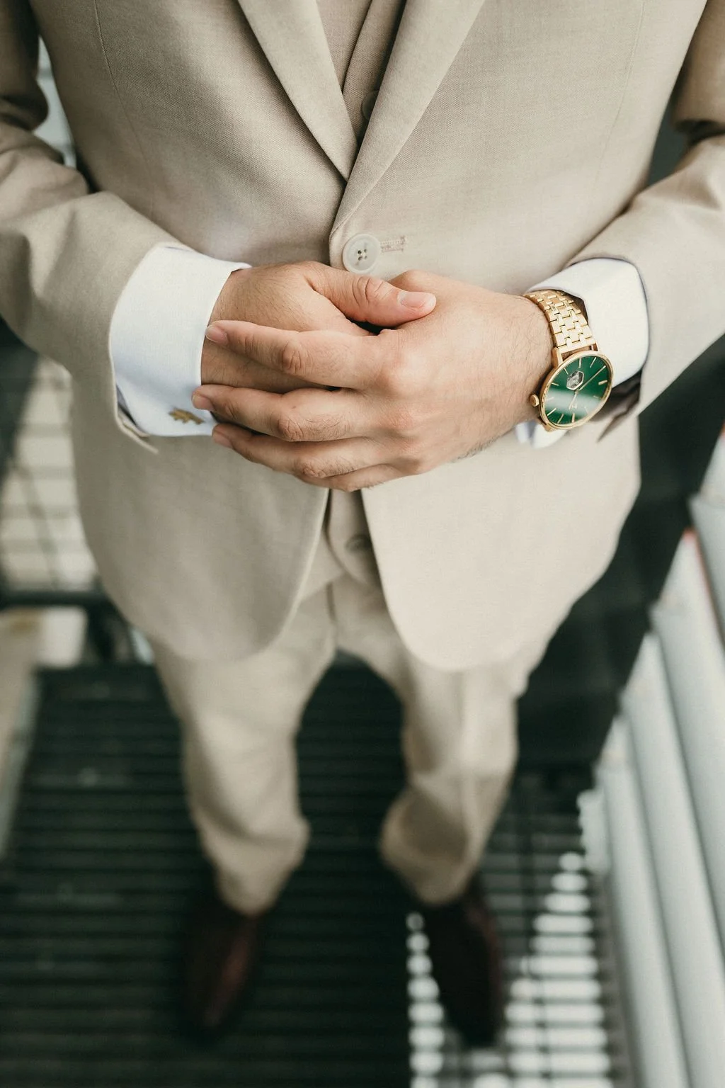 A man in a beige suit with a white shirt, wearing a gold watch with a green face, standing on a black metal walkway.