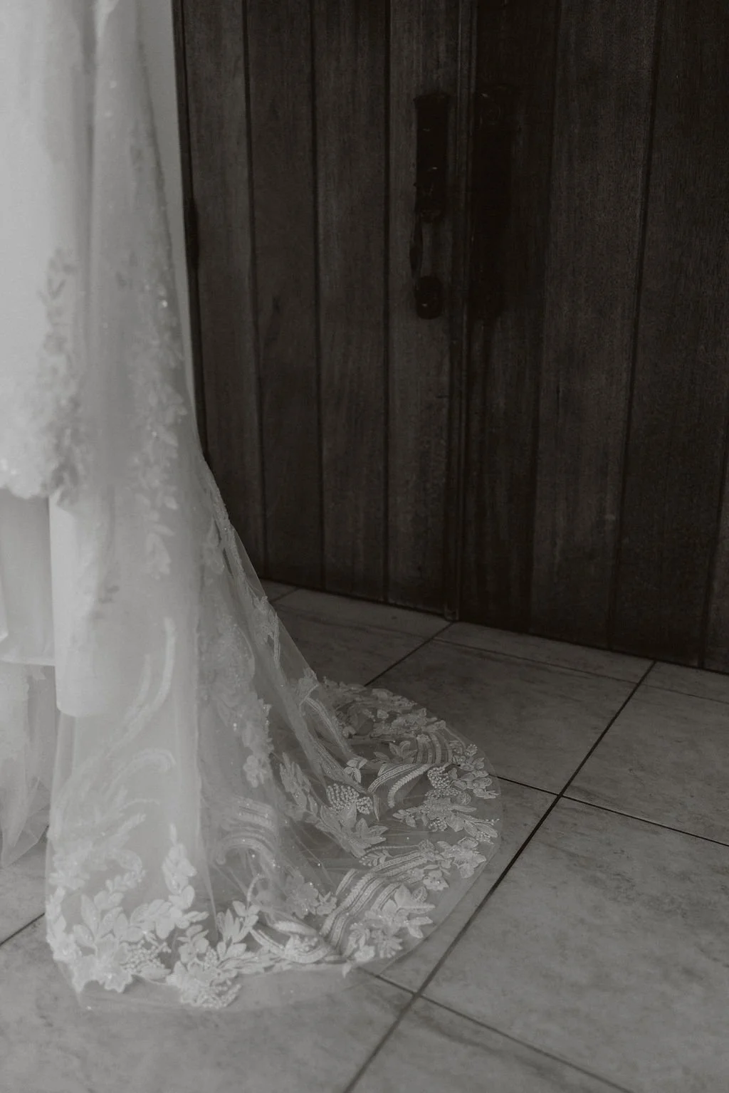 A close-up of a wedding dress train with lace details on the floor near a dark wooden door and tiled floor.