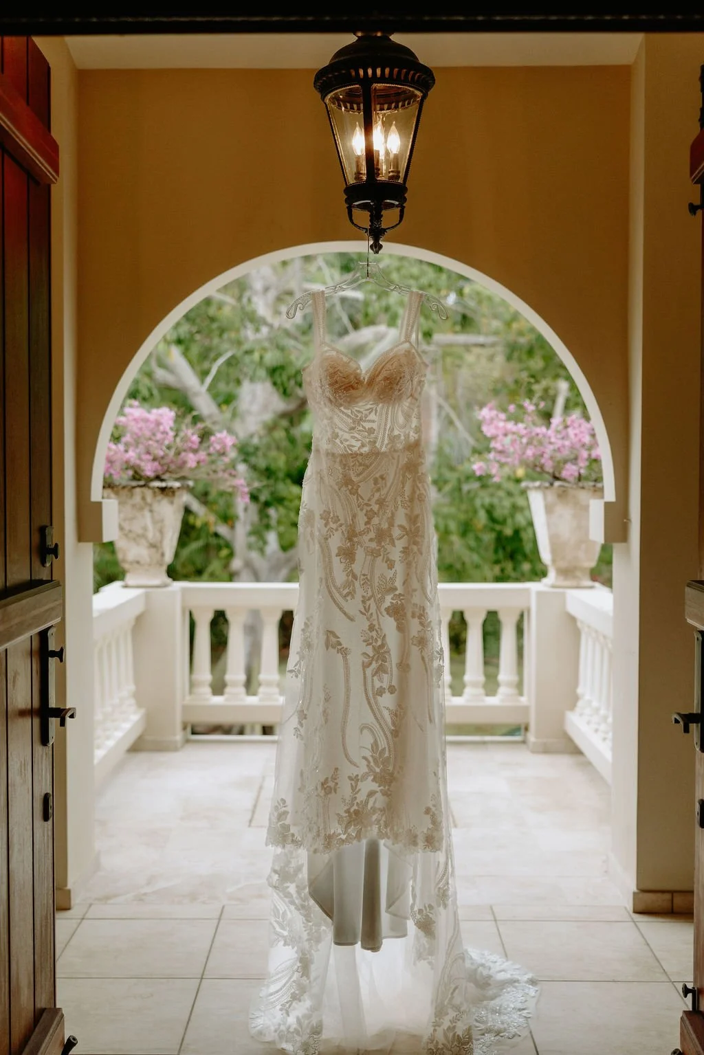 A wedding dress hanging from a lantern-like light fixture on a balcony with potted pink flowers and green trees in the background.