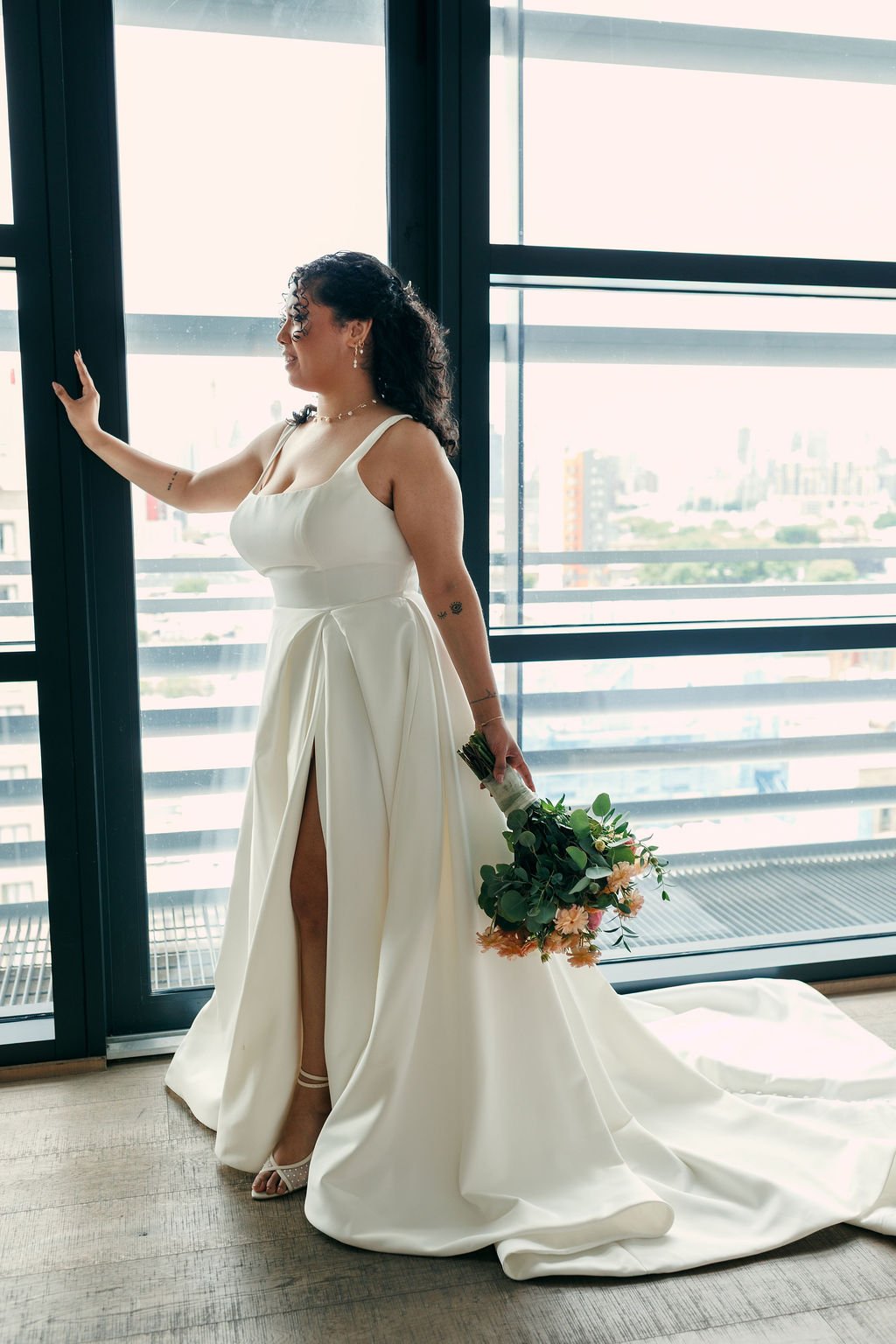 A bride in a white wedding dress standing by large glass windows, holding a bouquet of flowers, looking outside, with city buildings in the background.
