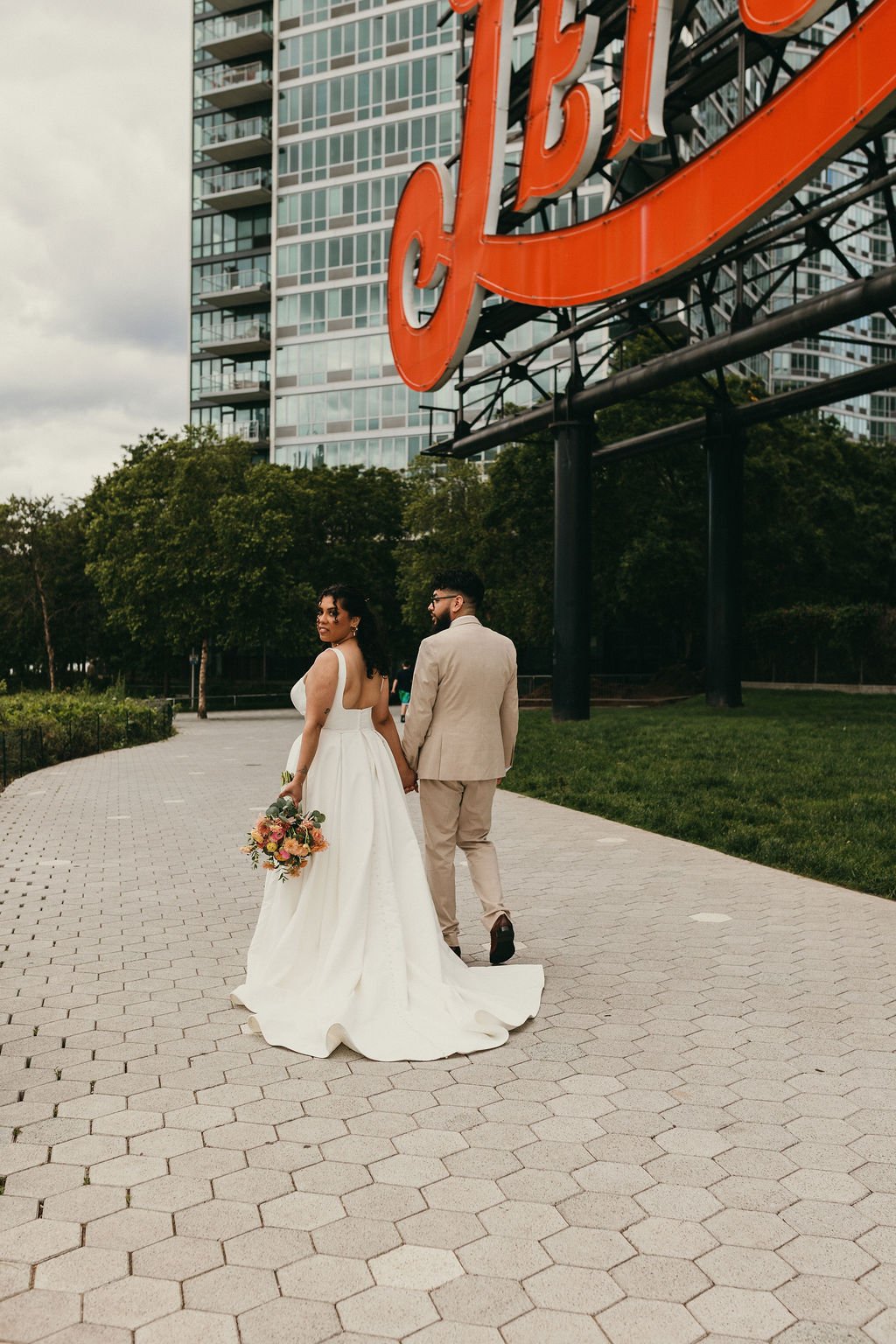 A bride and groom in wedding attire holding hands and walking away from the camera in an urban park, with a large orange sign and tall modern apartment buildings in the background.