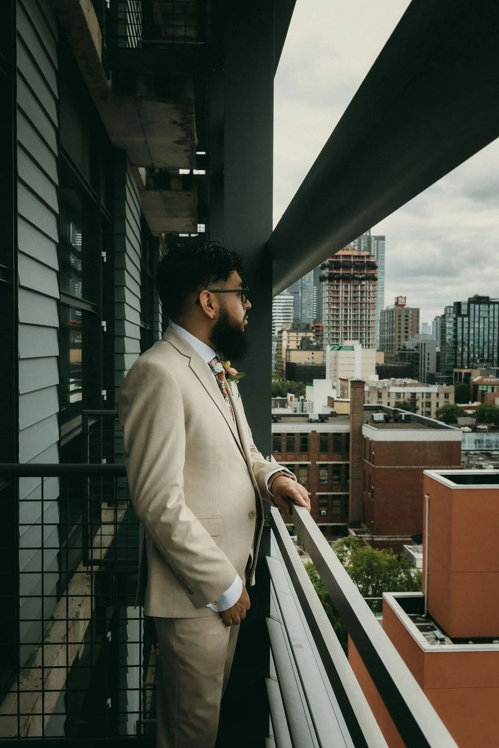 A man with a beard, glasses, and dark curly hair, dressed in a beige suit, stands on a balcony overlooking a cityscape with tall buildings under a cloudy sky.