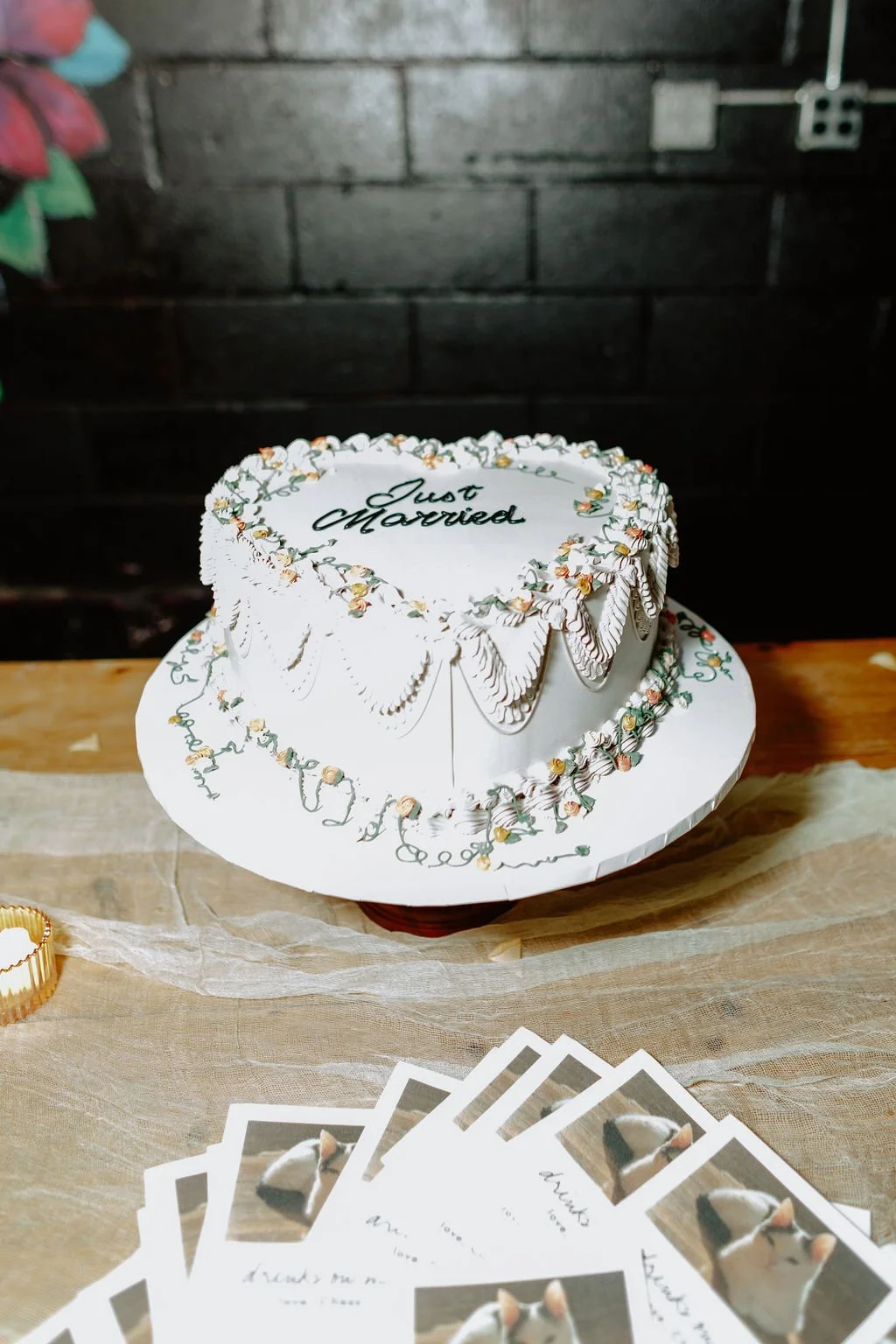 Heart-shaped wedding cake with white icing, decorated with a floral border, and the words 'Just Married' written on top. The cake is on a stand, and there are wedding favors or cards with animal pictures in front of it.