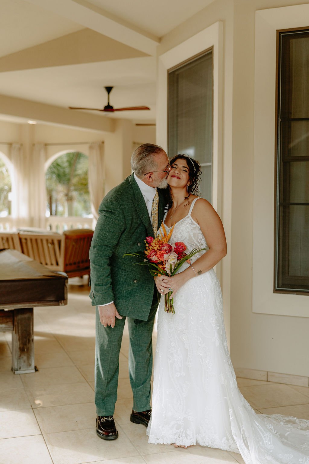 A bride in a white wedding dress holding a colorful bouquet is being kissed on the cheek by an older man in a green checkered suit inside a well-lit room with large windows and curtains.