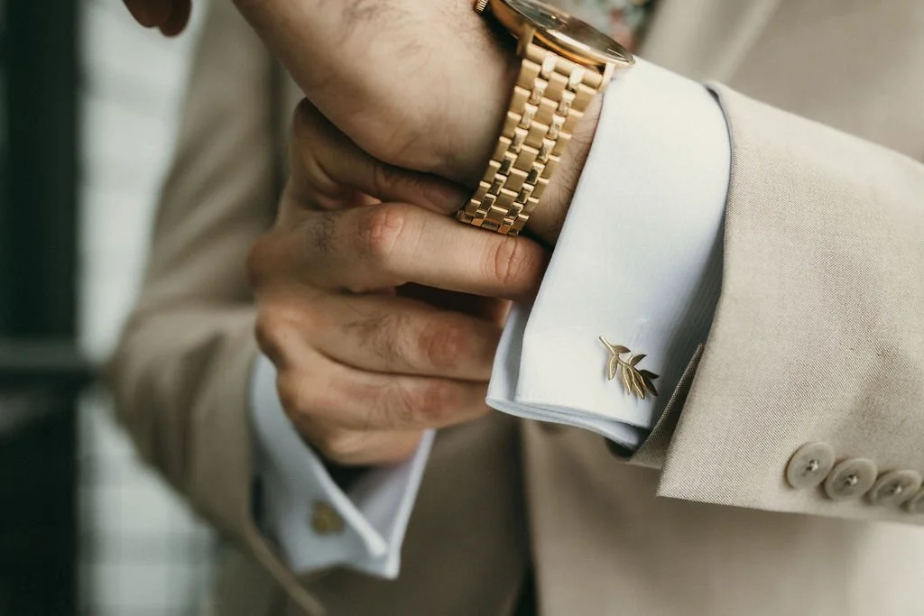 Close-up of a man's hand adjusting his gold wristwatch, wearing a beige suit jacket with a white shirt and a gold leaf cufflink.