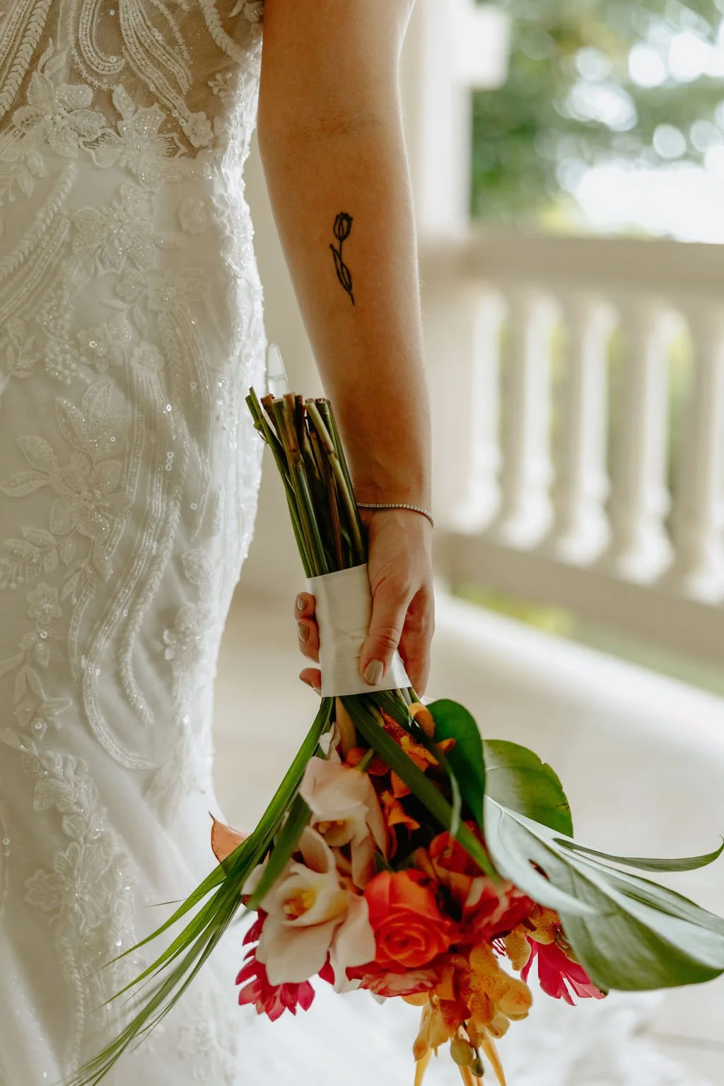 Close-up of a bride holding a colorful bouquet, part of her wedding dress visible, with a tattoo of a tulip on her arm, on a porch with a white railing and blurred greenery in the background.