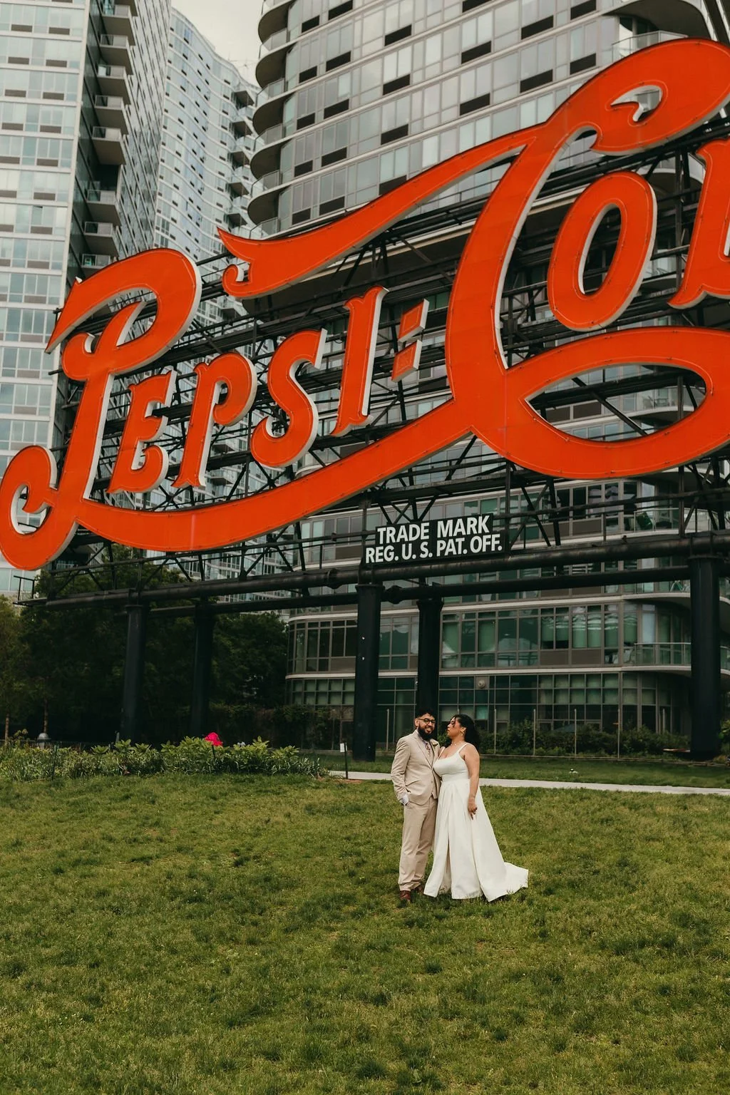 A couple in wedding attire standing on grass in front of a large neon Pepsi sign with modern apartment buildings in the background.