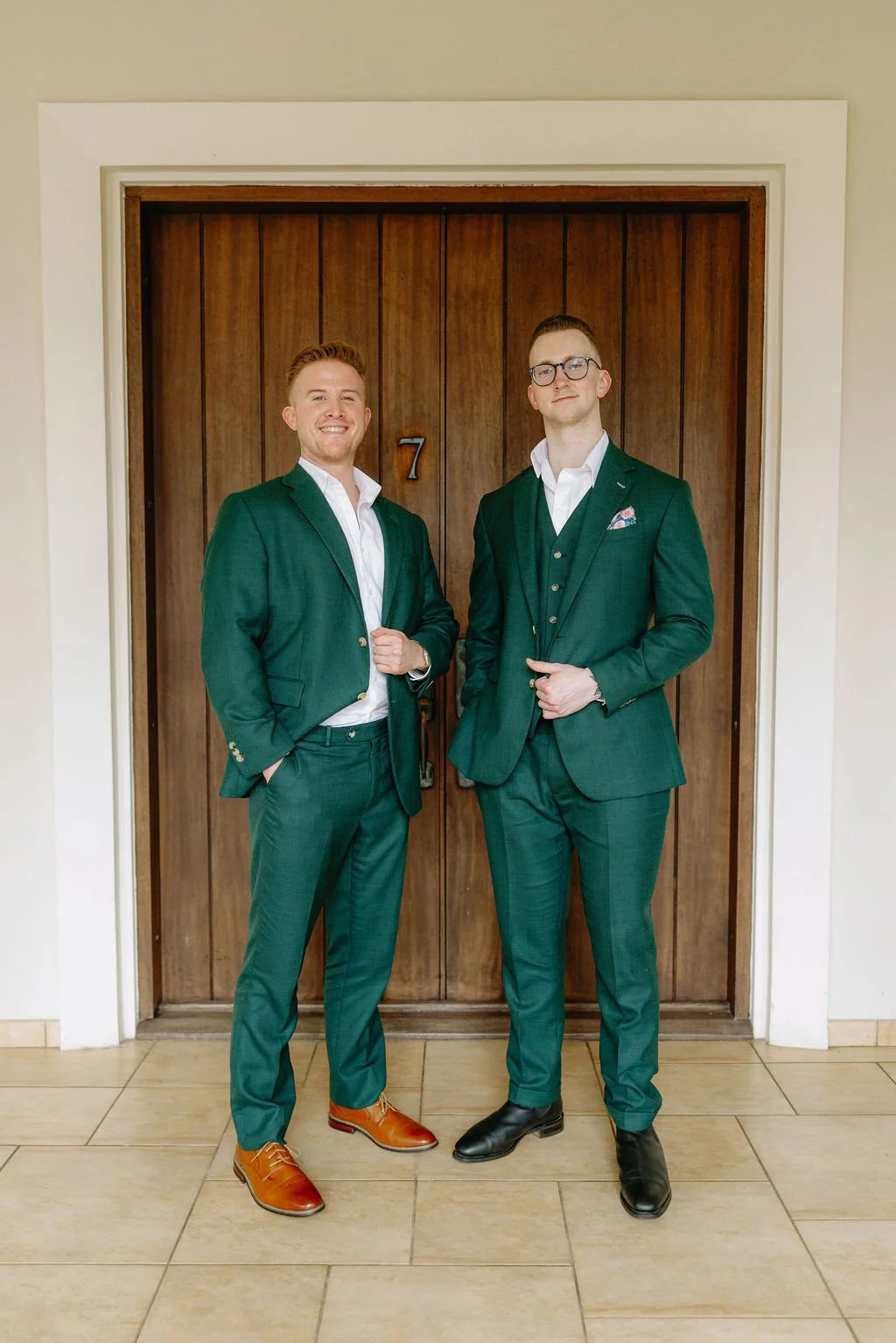 Two men dressed in green suits standing in front of a wooden door with the number 7, smiling and posing confidently.