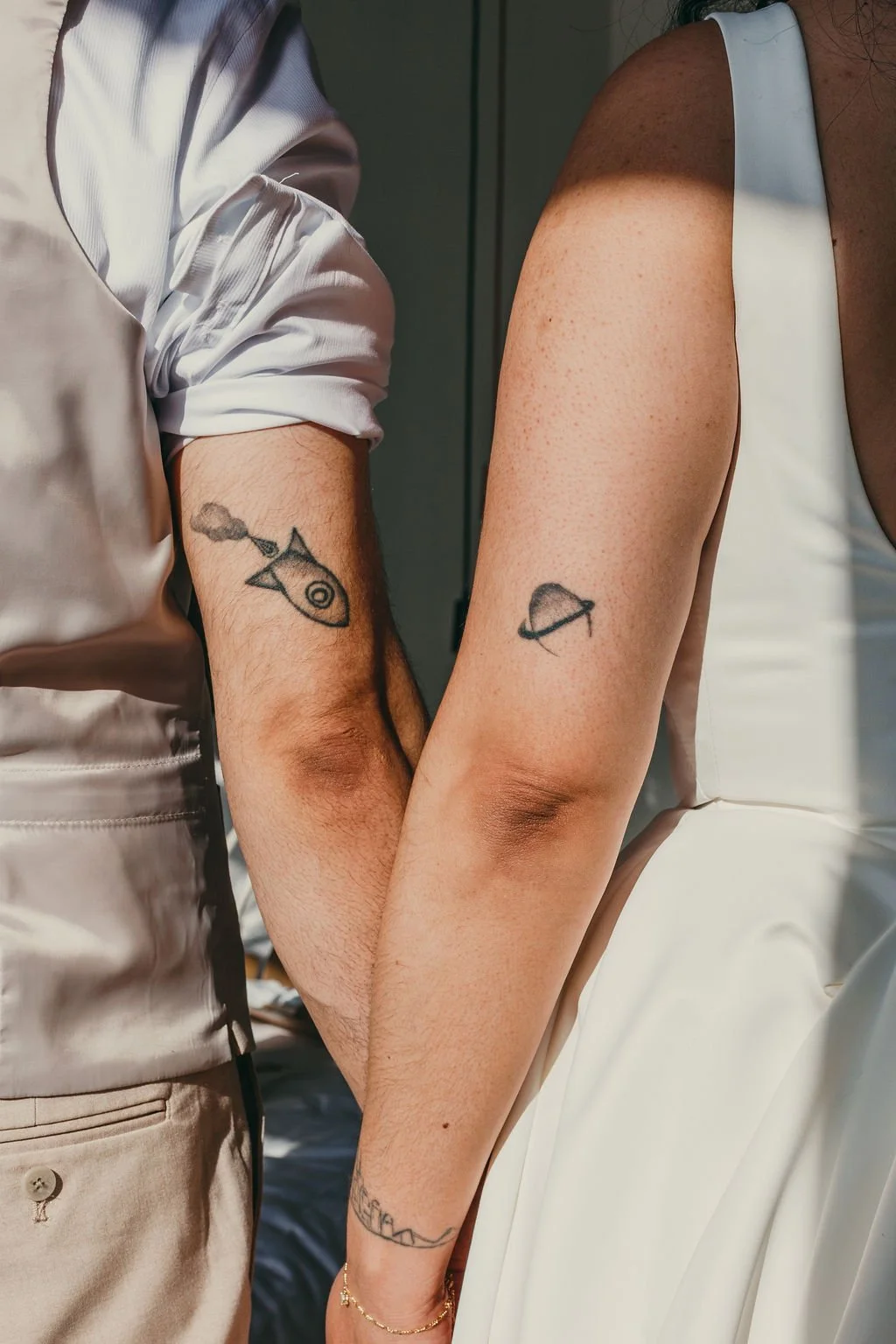 Close-up of a couple holding hands with space-themed tattoos: a spaceship on the male's arm and a planet with rings on the female's arm.