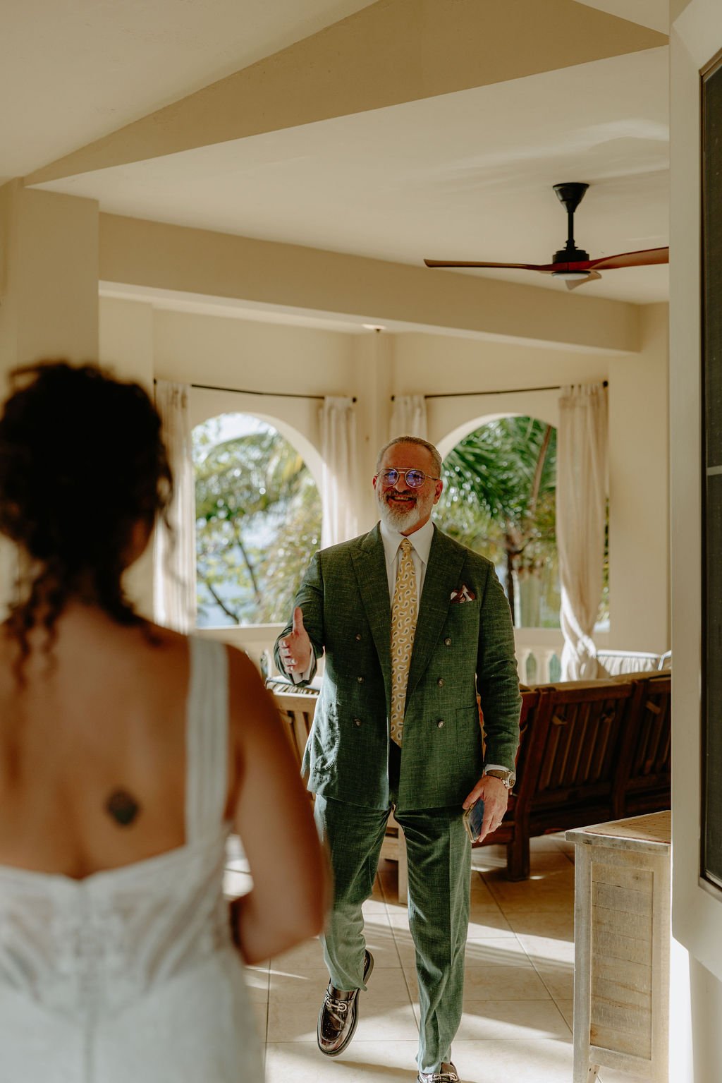 A man in a suit smiling and gesturing with his hand, standing in a bright room with large windows and curtains, with a woman in a white dress facing him.