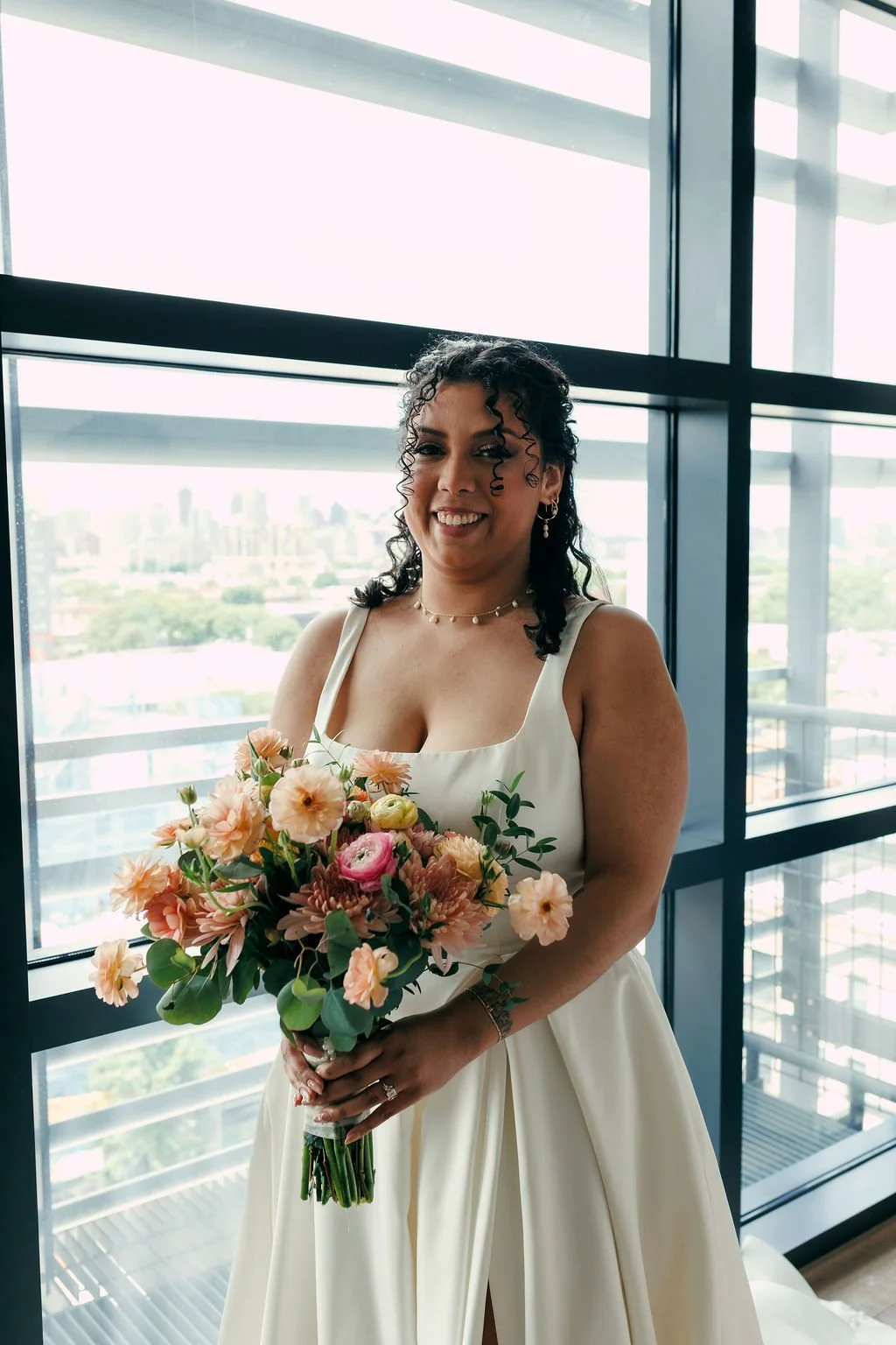 A woman in a white dress holding a bouquet of pink and peach flowers, standing indoors near large windows with a cityscape view in the background.