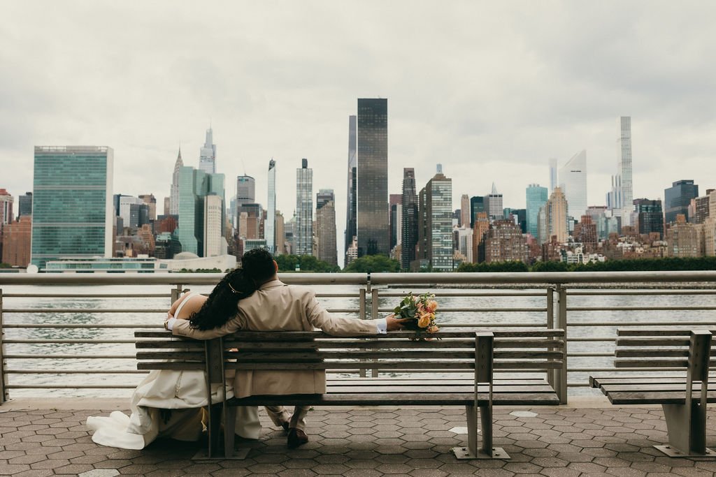A couple, possibly newlyweds, sitting on a bench by a river with a city skyline in the background, one holding a bouquet of flowers.