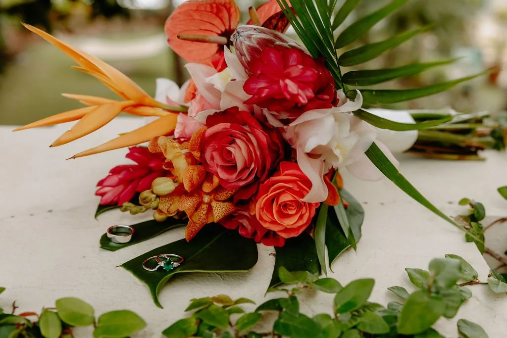 A colorful flower bouquet with roses, lilies, orchids, and tropical leaves, placed on a white surface with wedding rings underneath.