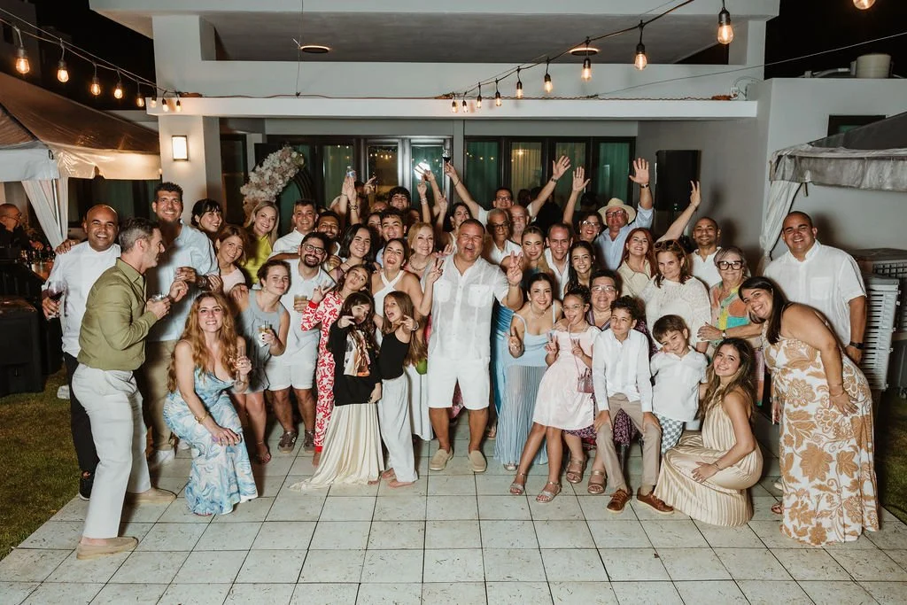 A large group of people gathered at a celebration at night, smiling and posing for photos on an outdoor patio with string lights overhead.