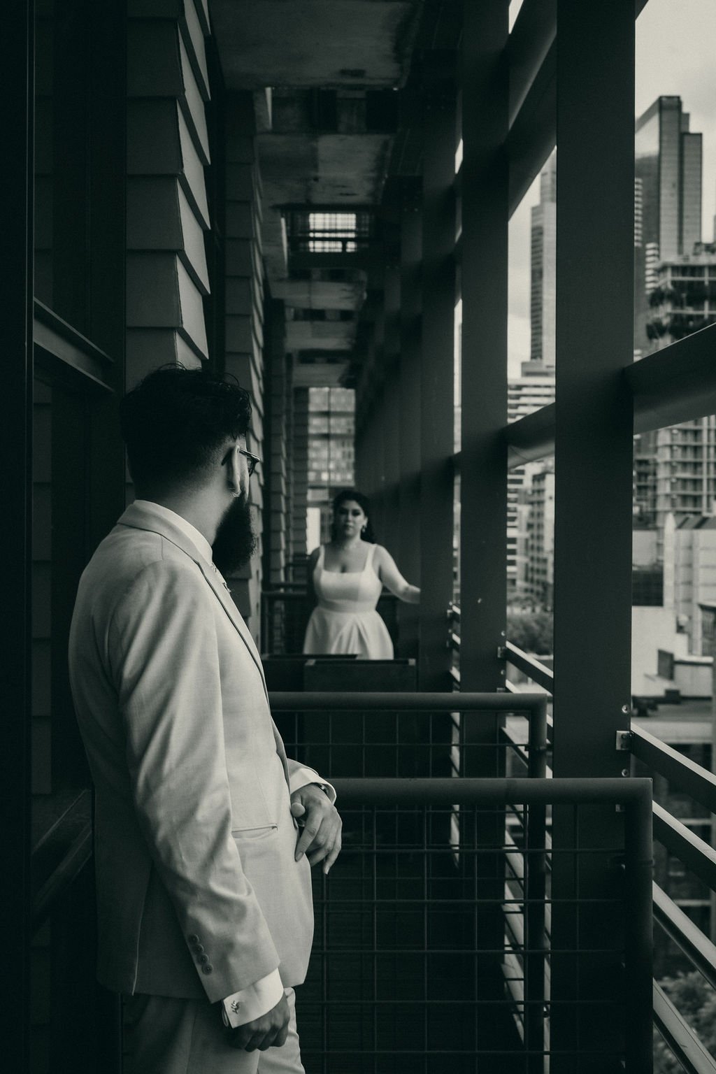A man in a light-colored suit with glasses and a beard looks at a woman on a balcony. The woman is wearing a sleeveless dress and is standing with her arm extended on the balcony railing. The shot is in black and white, and the background shows tall 