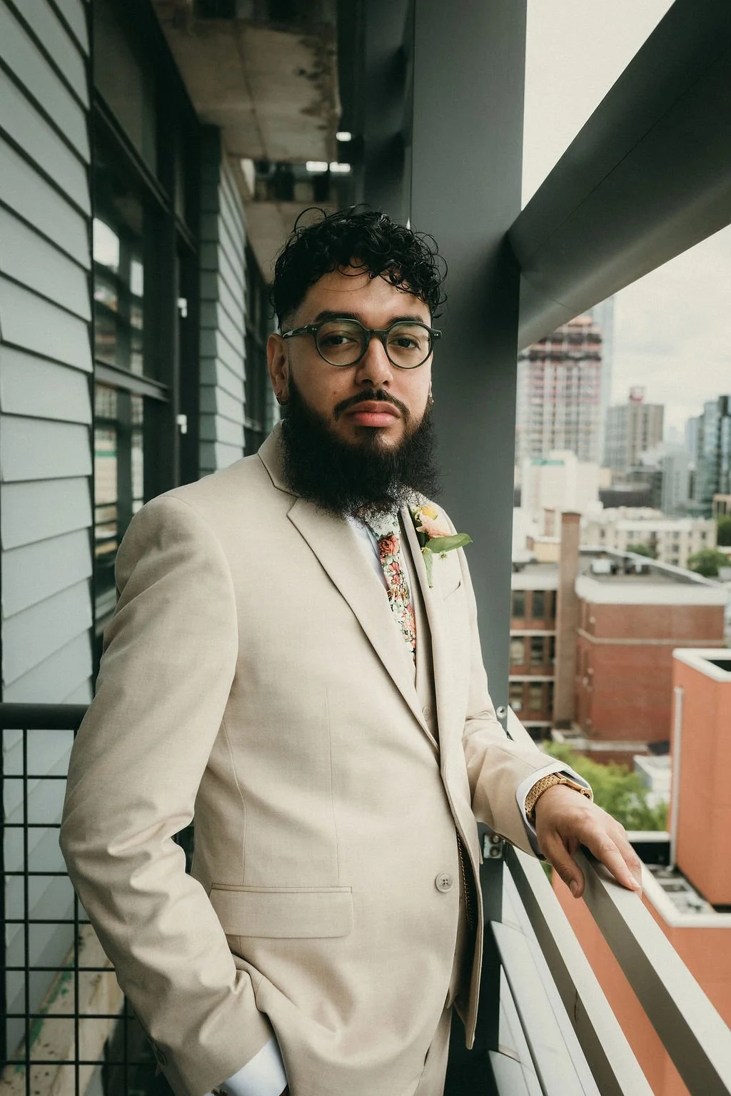 A man with curly hair and a beard wearing glasses and a beige suit standing on a balcony with a cityscape in the background.