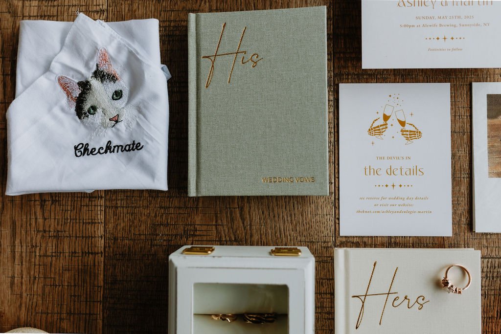 A wooden table displaying wedding-related items including a white T-shirt with a cat embroidery and the word "Checkmate," a green book titled "His Wedding Vows," a white card with gold accents about wedding details, a white book with the word "Hers" 