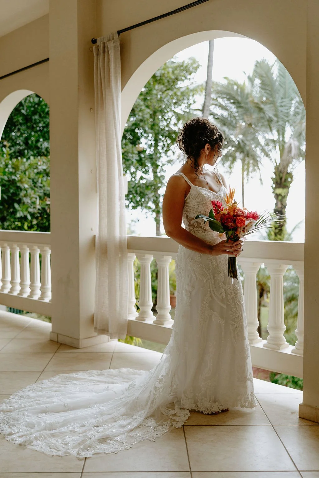 A bride in a white lace wedding dress holding a colorful bouquet of flowers, standing on a balcony with tropical trees in the background.