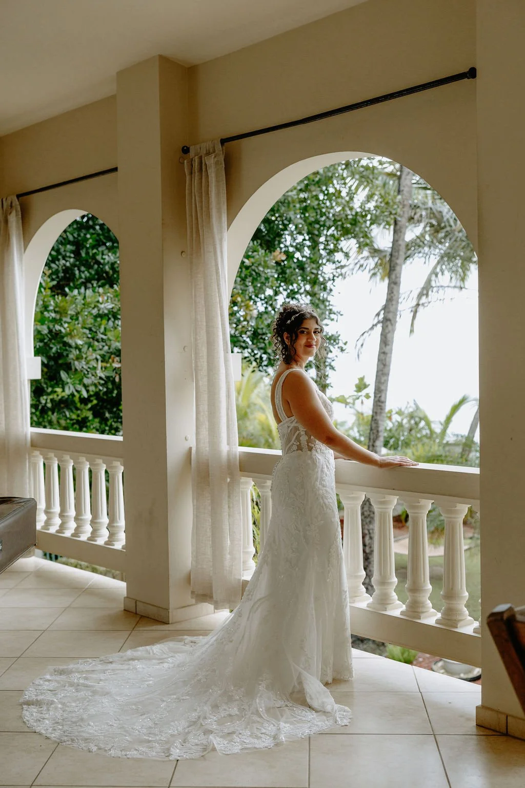A woman in a white wedding dress standing on a balcony with tropical trees outside.
