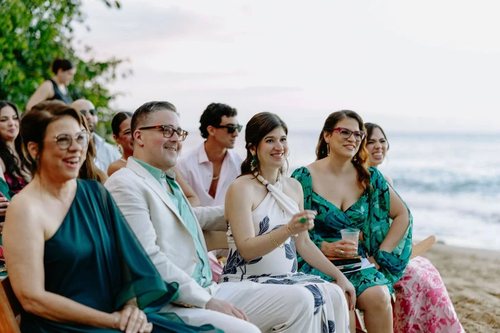 People in summer dresses and casual wear enjoying a beachside gathering or party, sitting on chairs near the ocean with trees in the background.