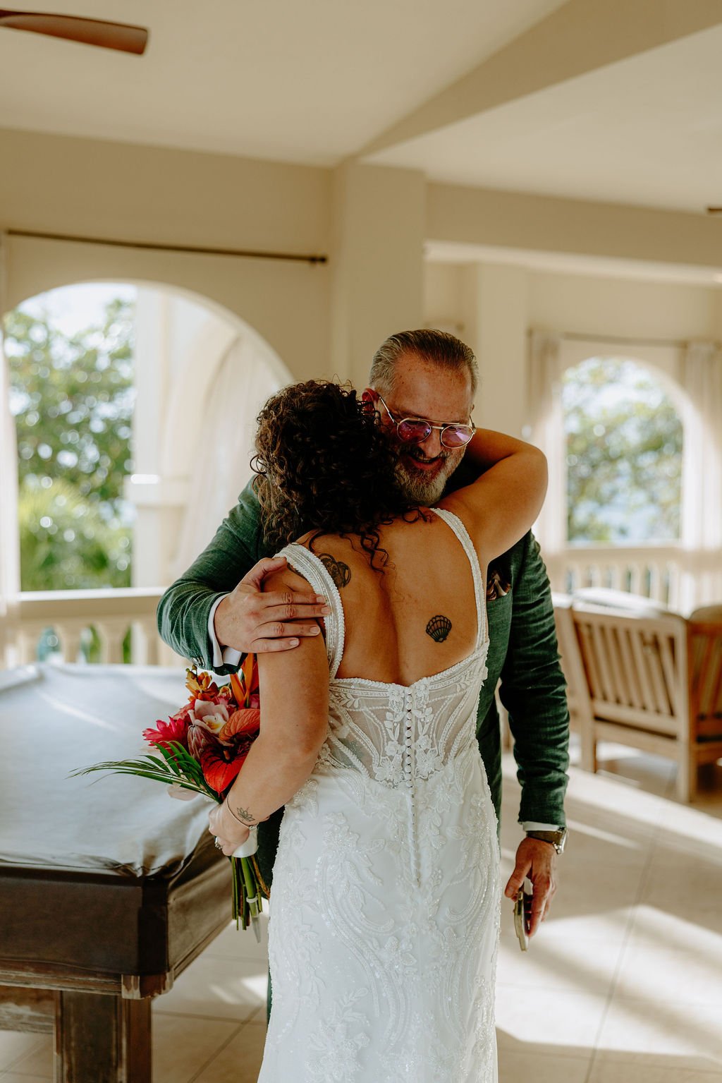 A man and woman are hugging each other, at a wedding or special occasion, indoors near large windows with natural light. The woman is holding a bouquet of flowers, and has a tattoo of a seashell on her back. The man is wearing glasses and a dark suit