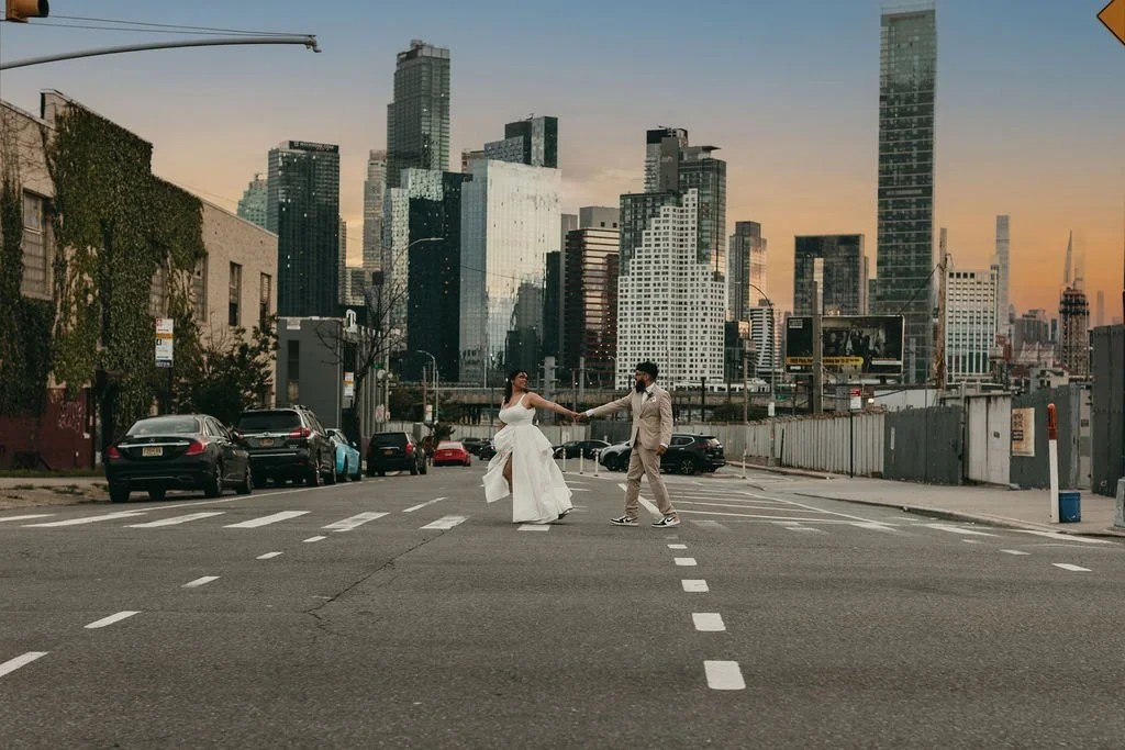 A couple in wedding attire holding hands while crossing a city street at sunset, with a skyline of tall skyscrapers in the background.