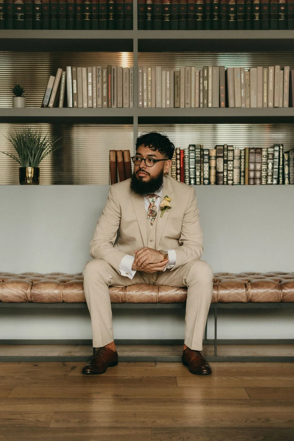 A man wearing a tan suit, glasses, and a floral tie, sitting on a tufted leather bench in front of a bookshelf with books and plants.