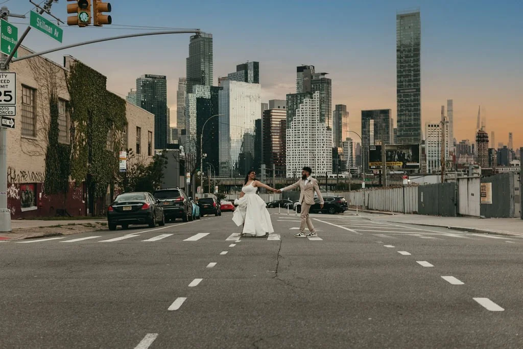 A couple dressed in wedding attire holding hands while crossing a city street intersection with skyscrapers in the background during sunset.