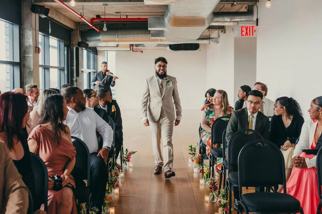 A man in a beige suit walking down an aisle at a wedding ceremony, with guests seated on either side and a violinist playing in the background.