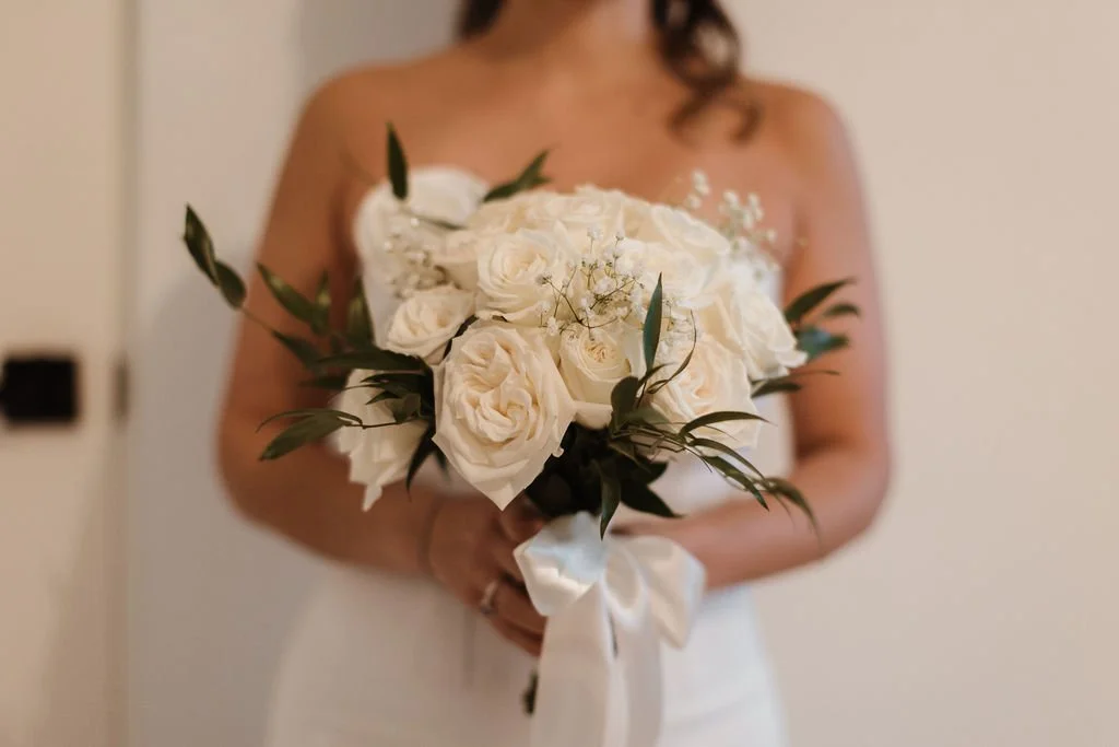 A woman in a white strapless dress holding a bouquet of white roses and greenery tied with a white ribbon.