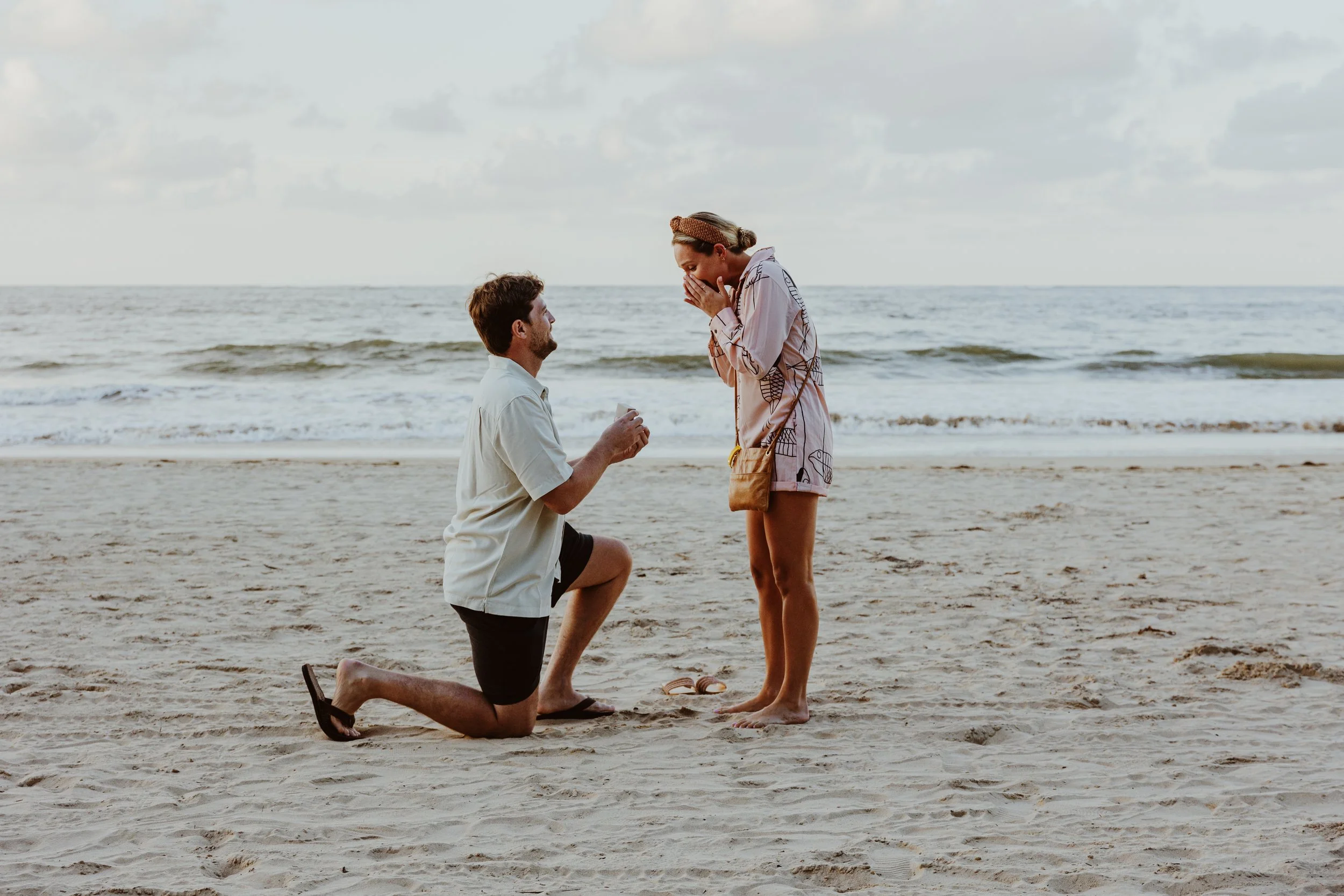 A man proposing marriage to a woman on the beach during sunset, with the man kneeling on one knee and holding a ring, and the woman covering her mouth in surprise.