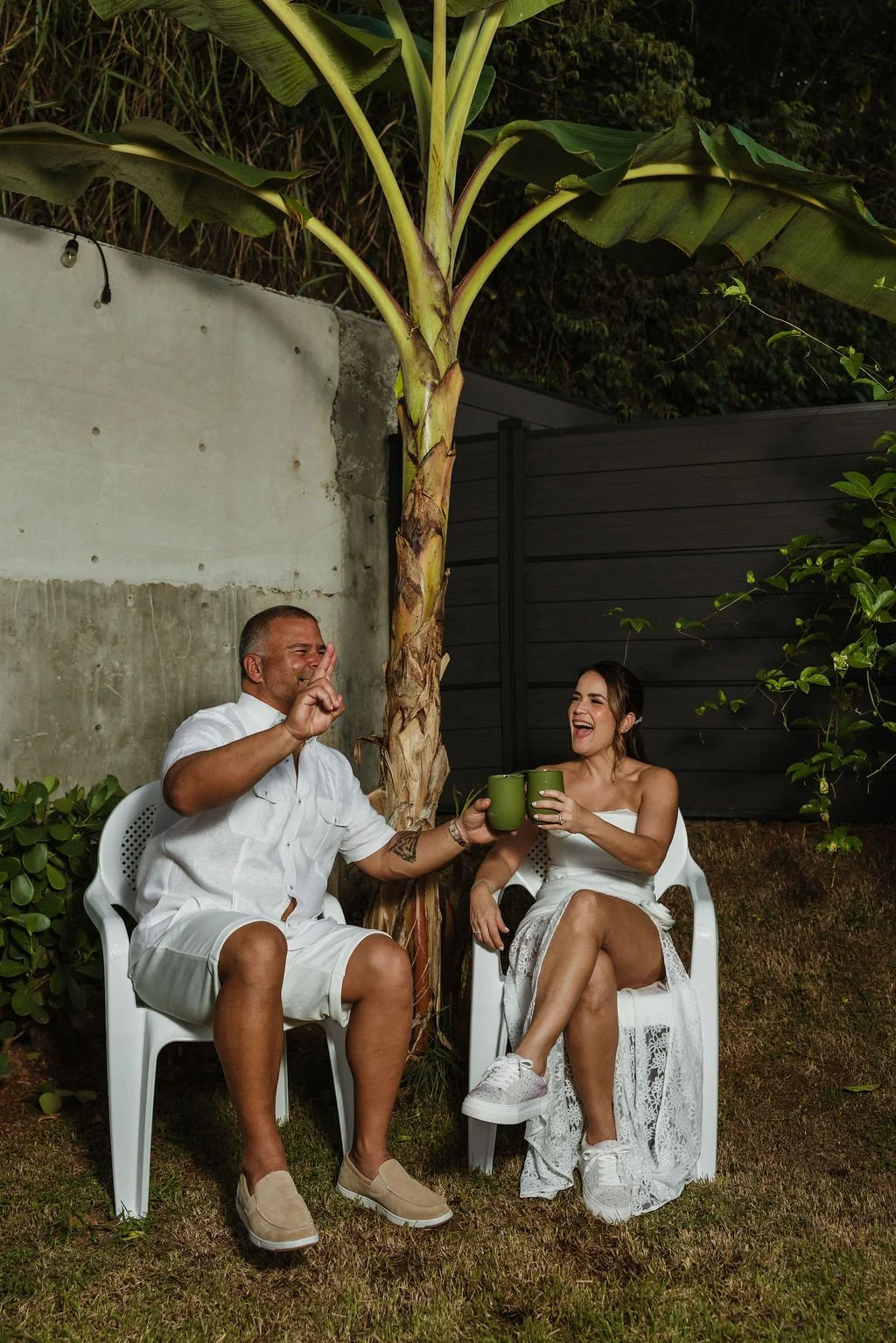 Two people sit outdoors under a large banana tree, laughing and holding green mugs, with a concrete wall and dark fence behind them.