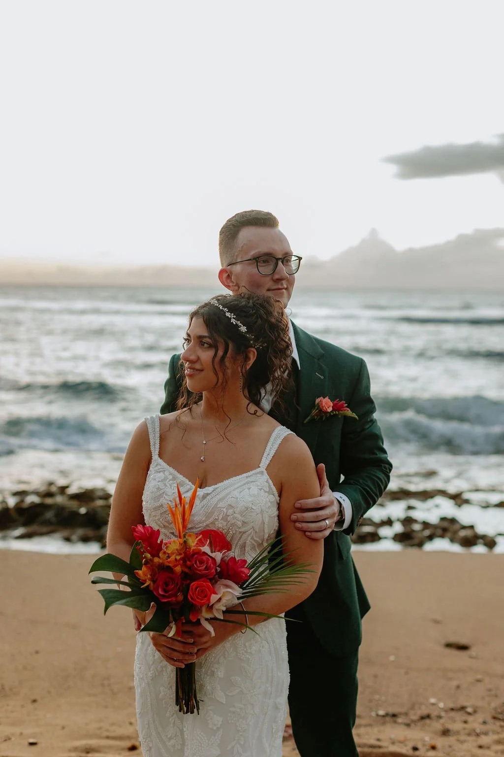 A bride and groom standing on a beach during sunset, with ocean waves in the background. The bride is holding a colorful bouquet and wearing a lace wedding dress, while the groom is dressed in a dark suit with a pink boutonniere.